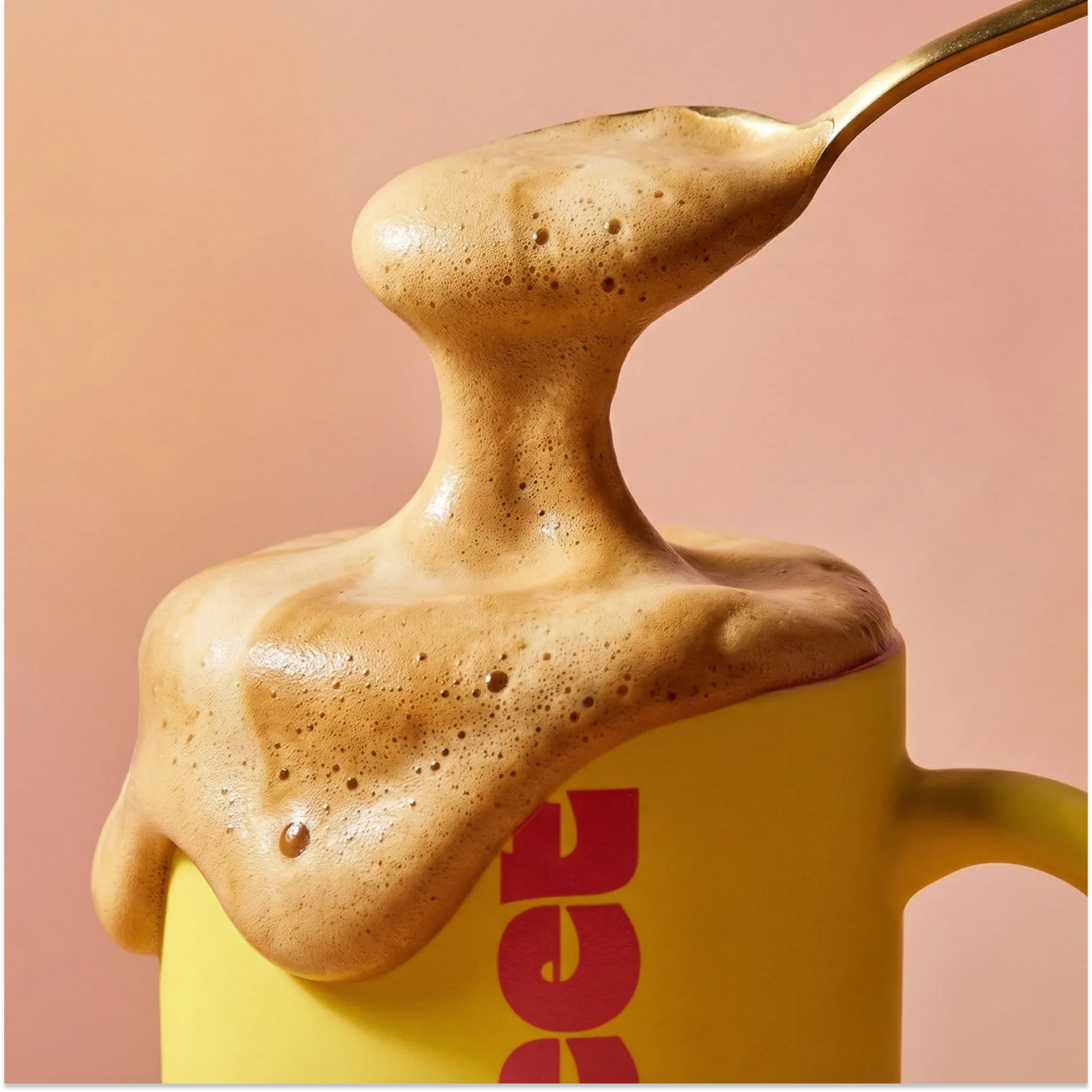 Thick coffee foam lifted on a spoon from a yellow SWEET mug against a pink background