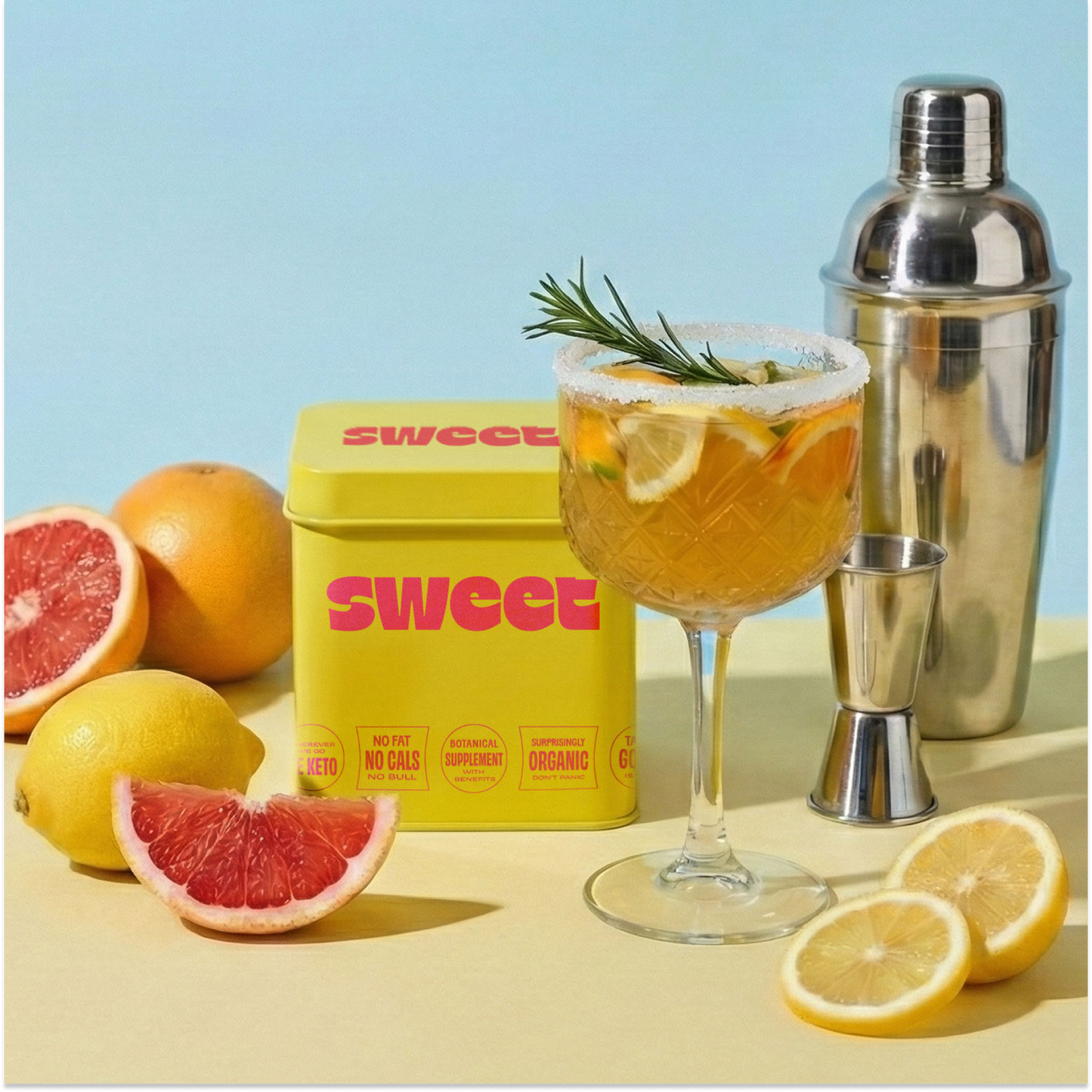 SWEET tin beside a citrus cocktail with rosemary, a shaker and halved grapefruit and lemon