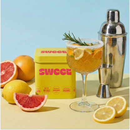 SWEET tin beside a citrus cocktail with rosemary, a shaker and halved grapefruit and lemon