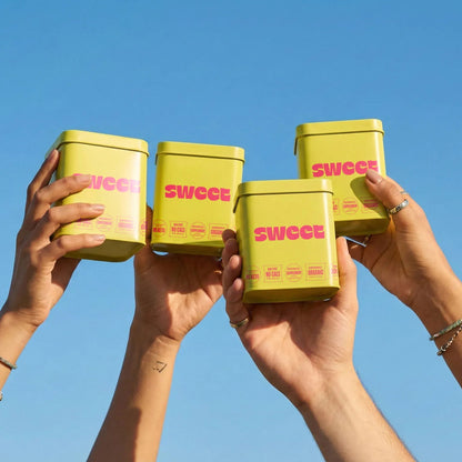 Hands raising SWEET square tins together in a toast against a blue sky