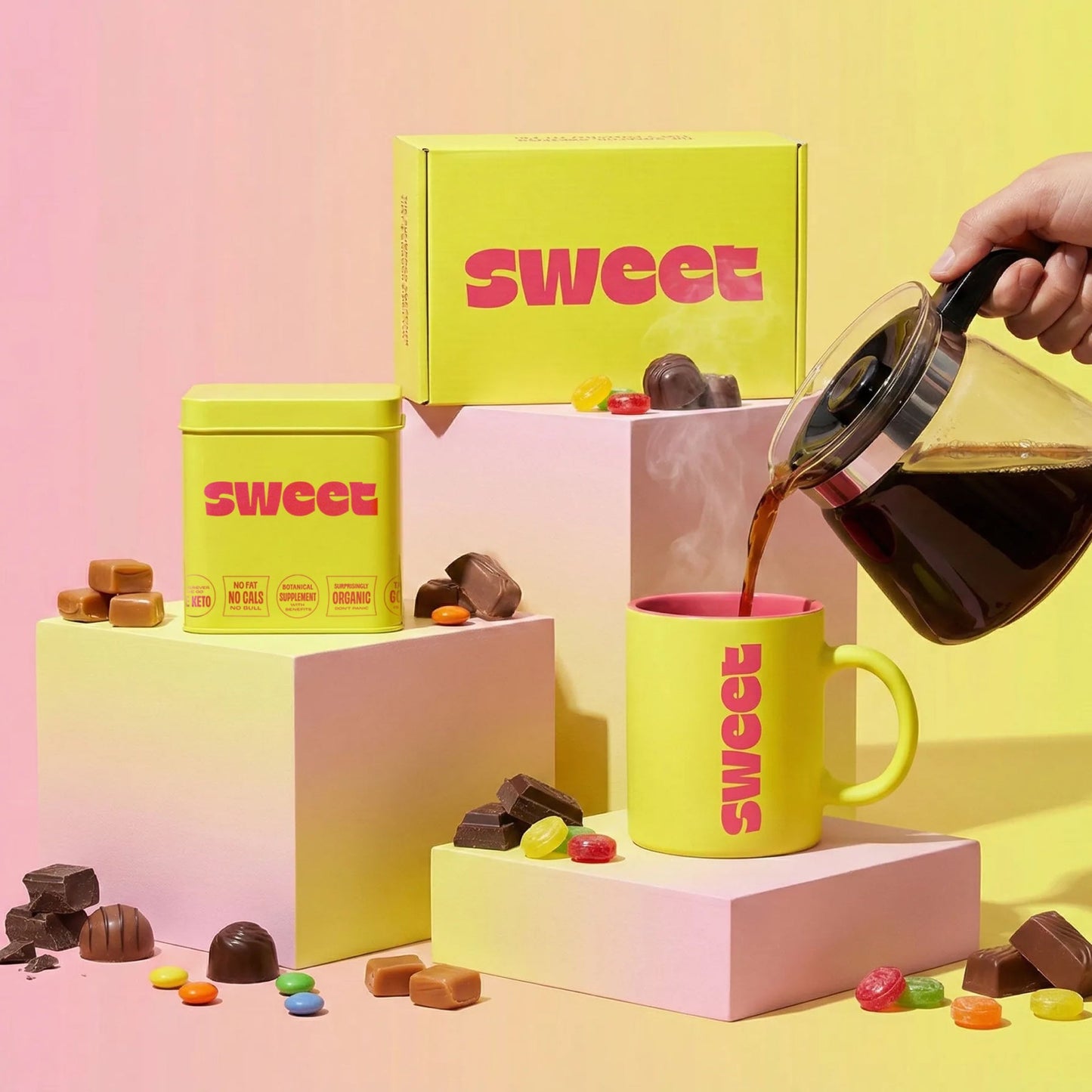 THE SWEETUAL™ — tin, mug, and box on pedestals with coffee poured from a carafe amid scattered chocolates and candies
