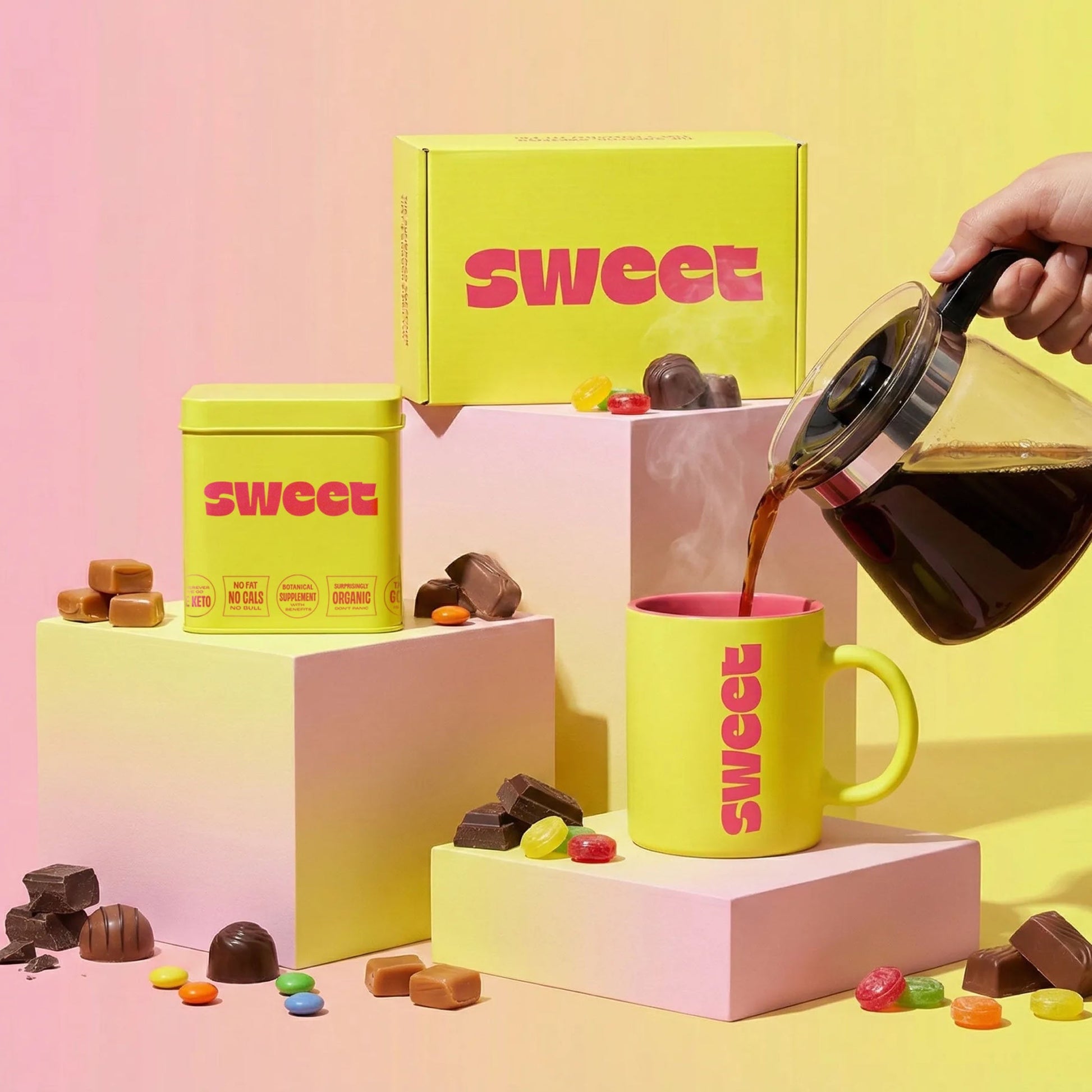 THE SWEETUAL™ — tin, mug, and box on pedestals with coffee poured from a carafe amid scattered chocolates and candies
