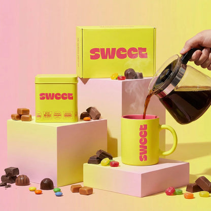 THE SWEETUAL™ — tin, mug, and box on pedestals with coffee poured from a carafe amid scattered chocolates and candies