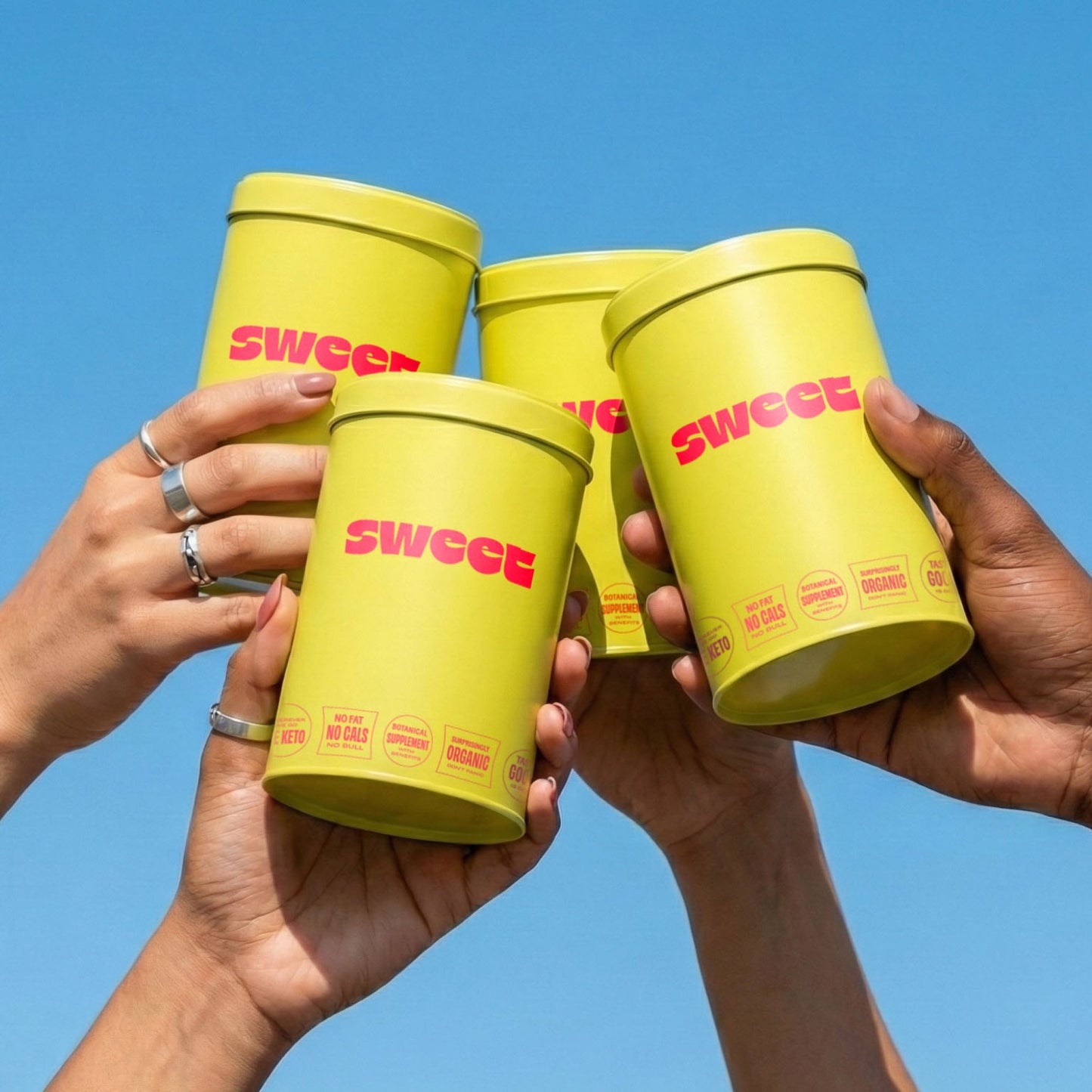 Hands raising SWEET tins together in a toast against a blue sky